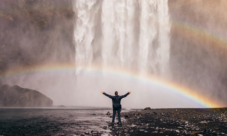 The Legacy Of Trauma And Positive Change man standing triumphant in front of waterfall and rainbow