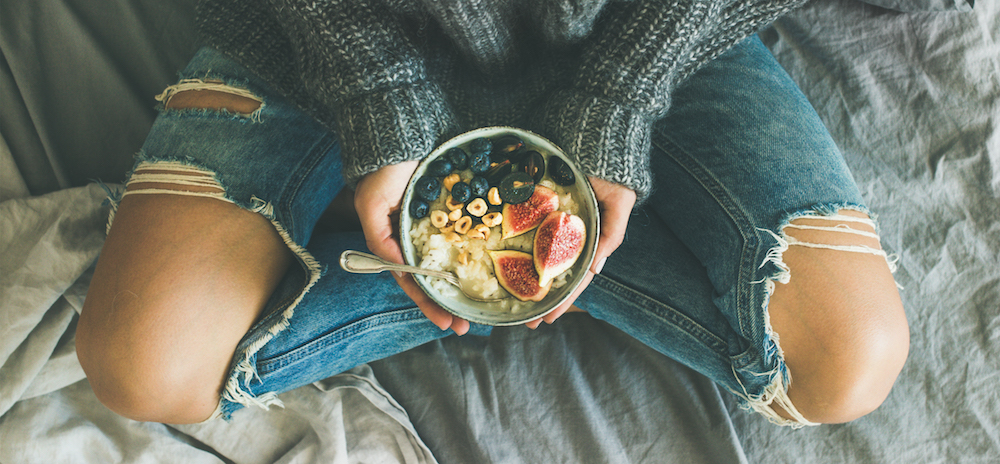 Superfood collagen. Healthy winter breakfast in bed. Woman in sweater and jeans holding rice coconut porridge with figs, berries, hazelnuts, top view, wide composition. Clean eating, vegetarian, comfort food concept